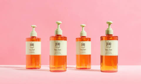 Four Pixi Glow Tonic pump bottles filled with orange liquid, arranged in a row against a solid pink background. View 1