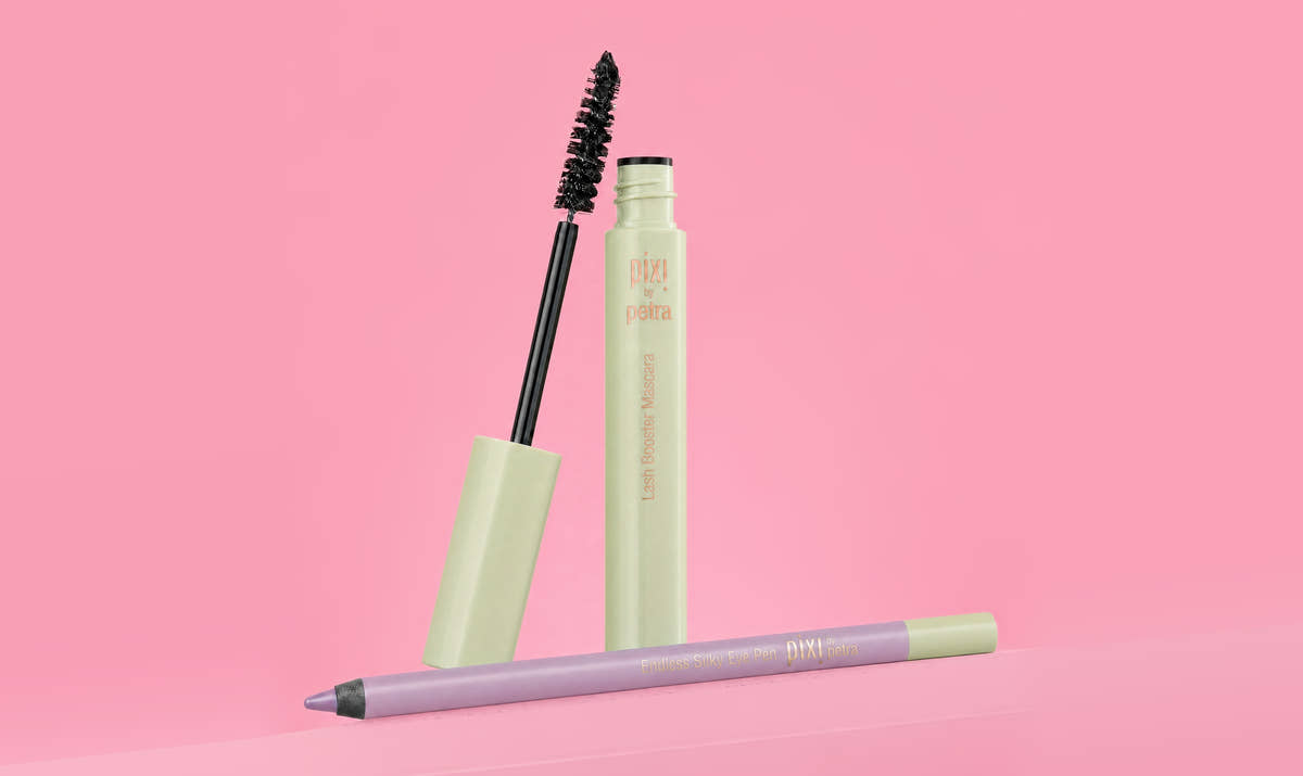 A cosmetic makeup set featuring a light green mascara tube with the wand removed and standing beside it, and a lavender lip liner pencil lying in front, all set against a solid pink background. View 1